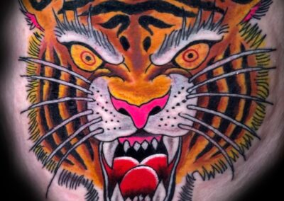 Bold American traditional tiger head tattoo with vibrant orange and black stripes, bright yellow eyes, pink nose, and open snarling mouth, expertly tattooed at Clockwork Tattoo Shop in Centereach, Long Island, New York.