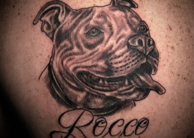 Black and grey realistic portrait tattoo of a smiling pit bull named Rocco with detailed shading and script lettering, tattooed at Clockwork Tattoo Shop in Centereach, Long Island, New York.