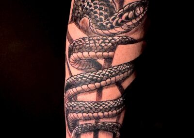 Black and grey snake wrapped around a dagger forearm tattoo with bold 24/8 lettering, featuring detailed scales and realistic shading, custom tattoo by Clockwork Tattoo Shop in Centereach, Long Island, NY.